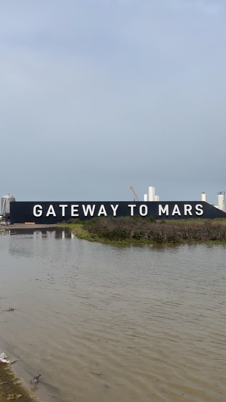 A long black wall carries the words GATEWAY TO MARS in tall white letters, seen across a wide stretch of shallow, still water and low scrub. Stainless tanks, an orange lattice crane boom and a domed stainless structure stand behind it under a flat grey sky, and a small shorebird stands in the shallows in the foreground.