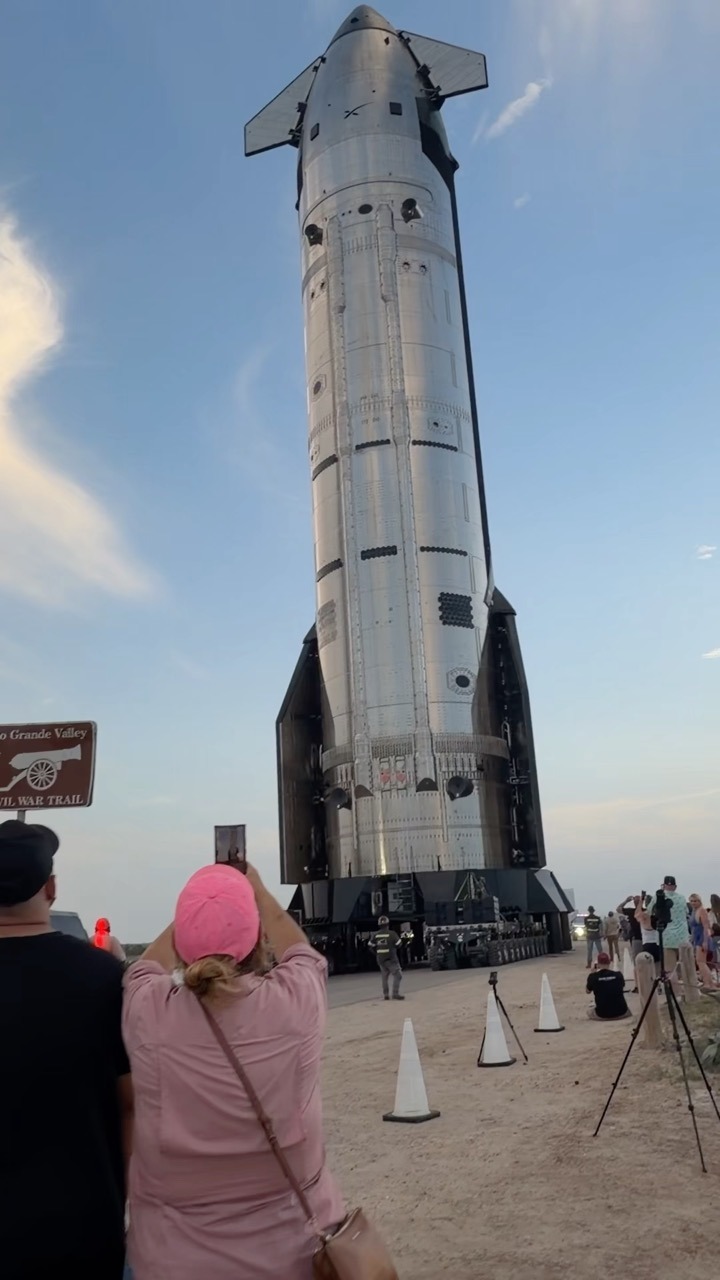 A stainless steel Starship upper stage rides upright on a low multi-wheeled transporter along a paved road with a sandy shoulder, towering over the crowd watching from the roadside. Spectators film it on phones and tripod-mounted cameras between traffic cones, and a brown roadside marker stands at the left of the frame.