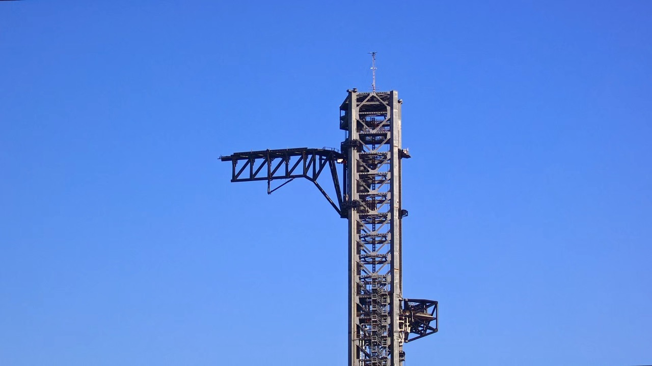 The upper section of a tall steel lattice launch tower against a clear deep-blue sky, with a long hinged arm extended out to one side and a lightning mast on top.