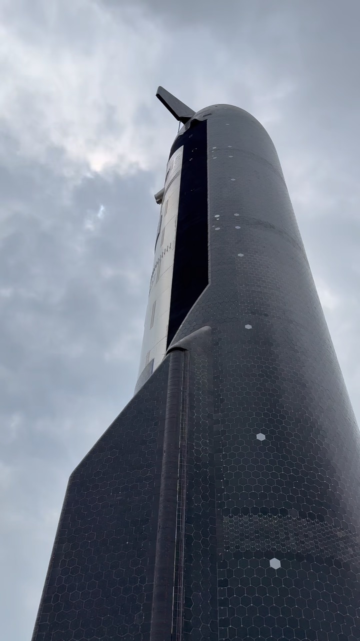 Looking steeply up the flank of a Starship upper stage, its hexagonal black heat-shield tiles filling the frame, a forward flap jutting out at the top against a grey overcast sky.