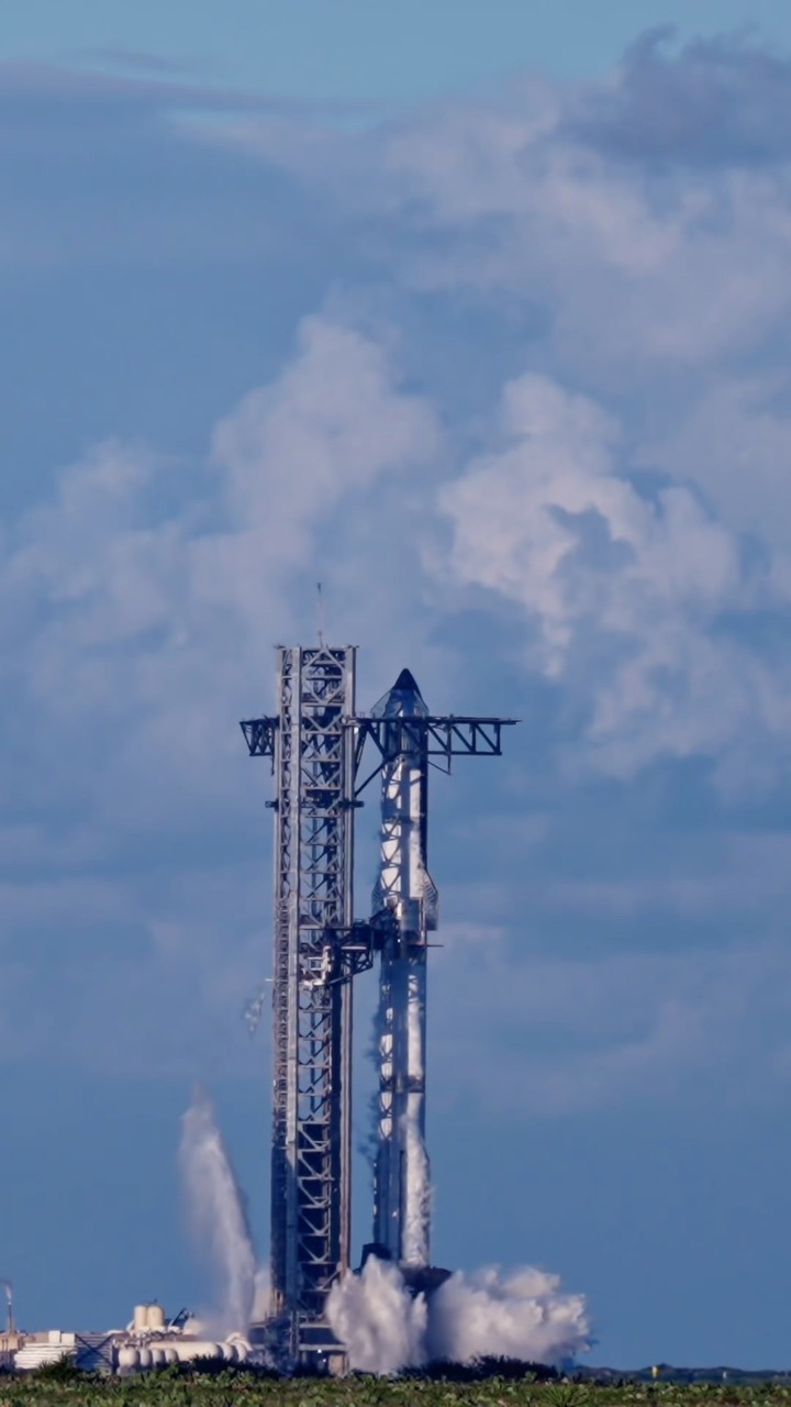A stacked Starship and Super Heavy stand on the pad beside the launch tower, venting vapour along the vehicle, with water spray rising at the base under a tall bank of cumulus.