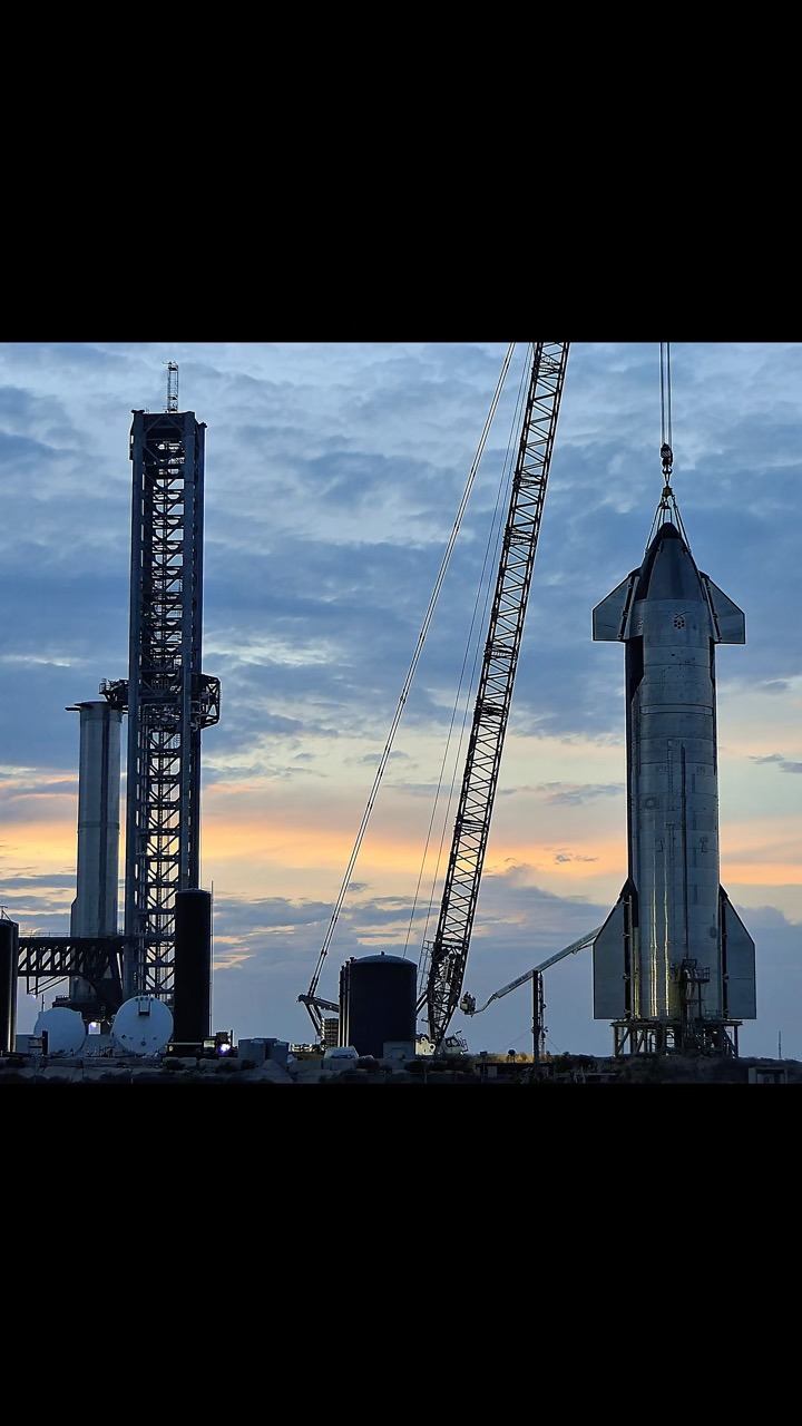 A Starship upper stage stands upright on a stand at dusk beside a tall steel lattice tower, a lattice-boom crane line running down to a fitting at its nose, with tanks and ground equipment along the horizon against orange and blue cloud.
