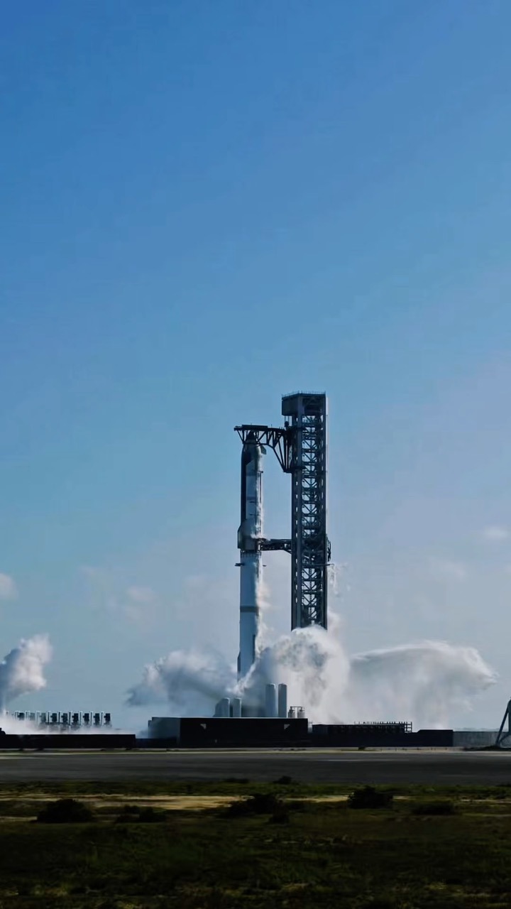 A stacked Starship and Super Heavy stand beside the launch tower, pale with frost down the tanks, with white vapour and spray billowing across the base of the pad, seen across a flat green plain under a clear blue sky.