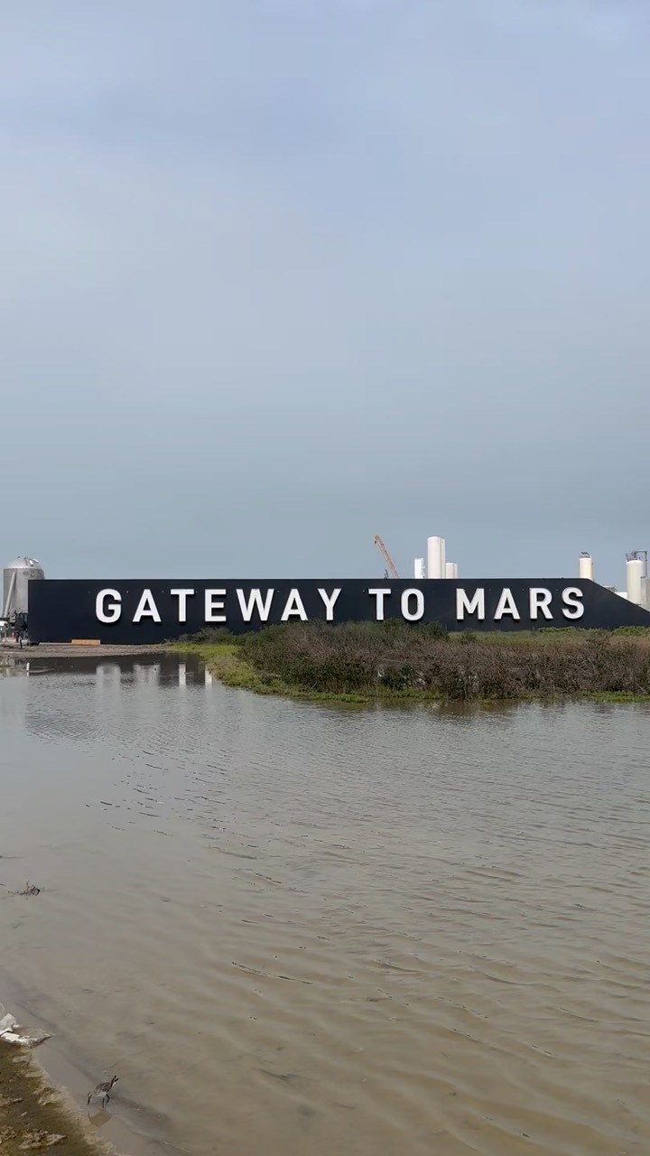 A long black wall lettered GATEWAY TO MARS stands beyond a flooded flat, with a wading bird in the shallow brown water, and white cylindrical tanks, an orange crane and a stainless vehicle section rising behind it.