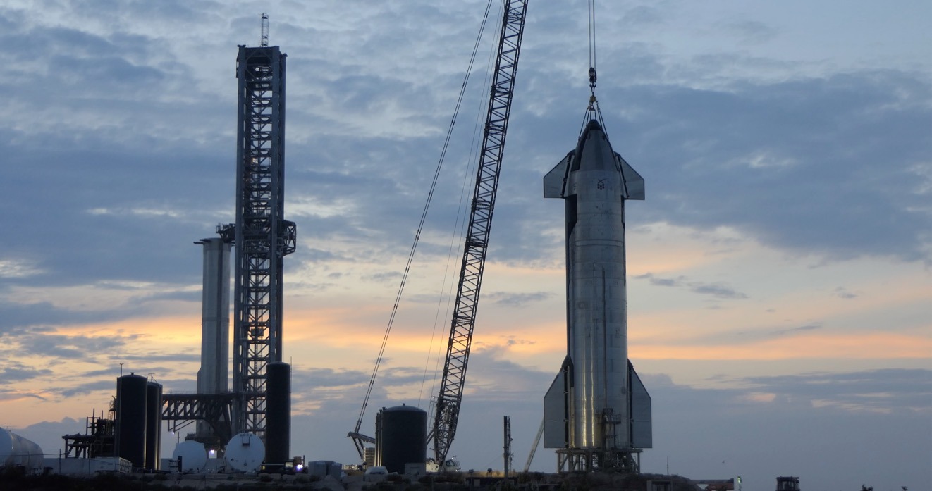 A stainless-steel Starship upper stage, its nose cone and four flaps visible, stands on a lattice support stand with a crane's hook and lifting slings rigged to its nose. A tall lattice tower, storage tanks and ground equipment stand nearby under a low, colour-streaked sky.
