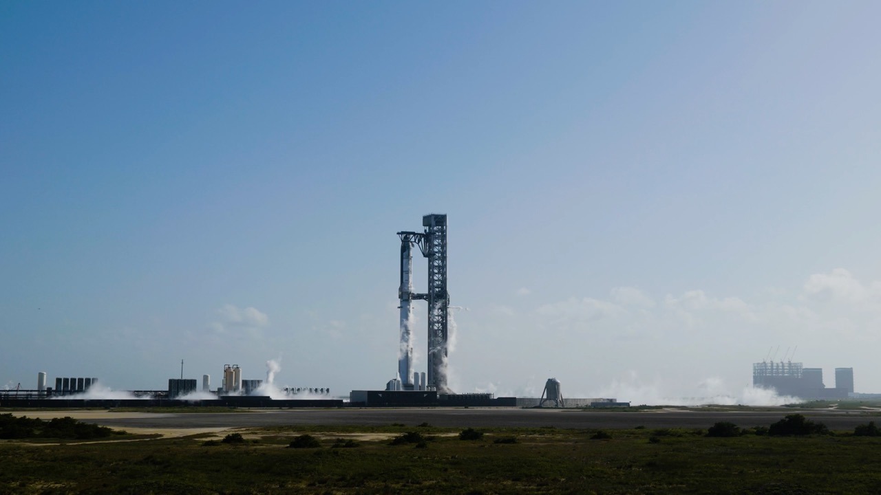 A stacked Starship and Super Heavy stand on a raised launch mount beside a tall tower with catch arms, white vapour venting along the base, seen from a distance across flat scrubland under a clear daylight sky.