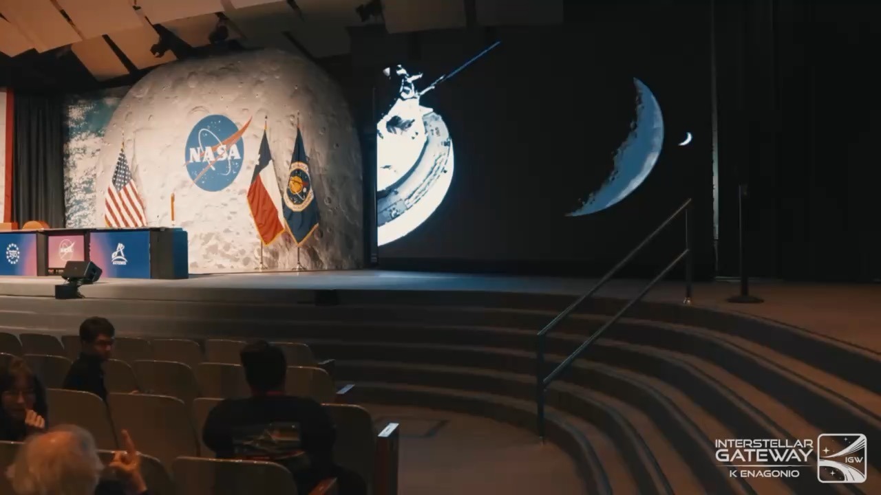 A briefing-room stage set against a large mural of the lunar surface carrying the NASA insignia, flanked by a US flag and two other flags, with a lectern table in front and screens beside the stage showing a spacecraft and a thin crescent Moon. A few people sit in the tiered auditorium seats in the foreground.
