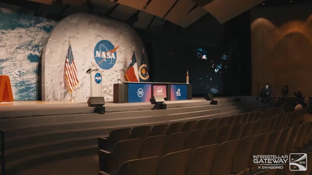 An auditorium stage with a lectern bearing the NASA insignia, set against a large mural of the lunar surface with the same insignia, a US flag and two other flags behind it, a small rocket model on the table and a dark screen to the side showing spacecraft imagery. Rows of empty seats fill the foreground.