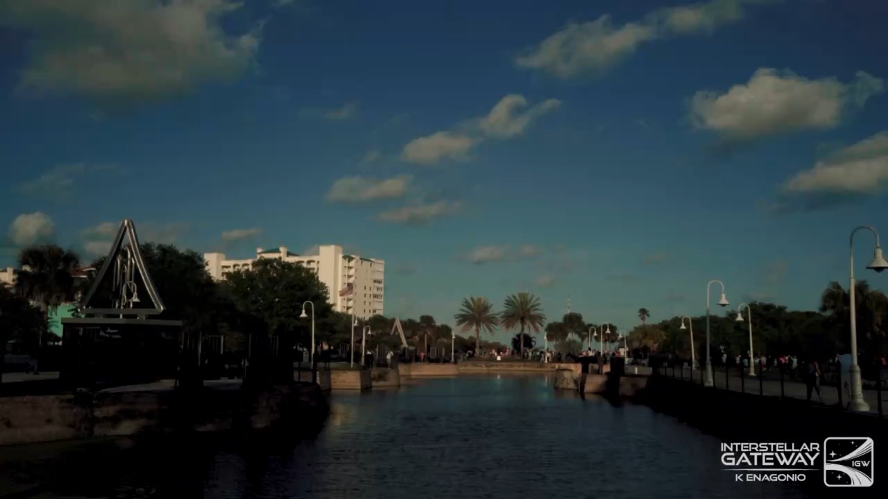 A calm waterfront basin under a blue sky with broken cloud, its stone-walled banks lined with street lamps and palm trees. A white mid-rise building and a tall triangular metal structure stand beyond the banks, a scattered crowd is gathered along the far side, and the foreground is in deep shade.