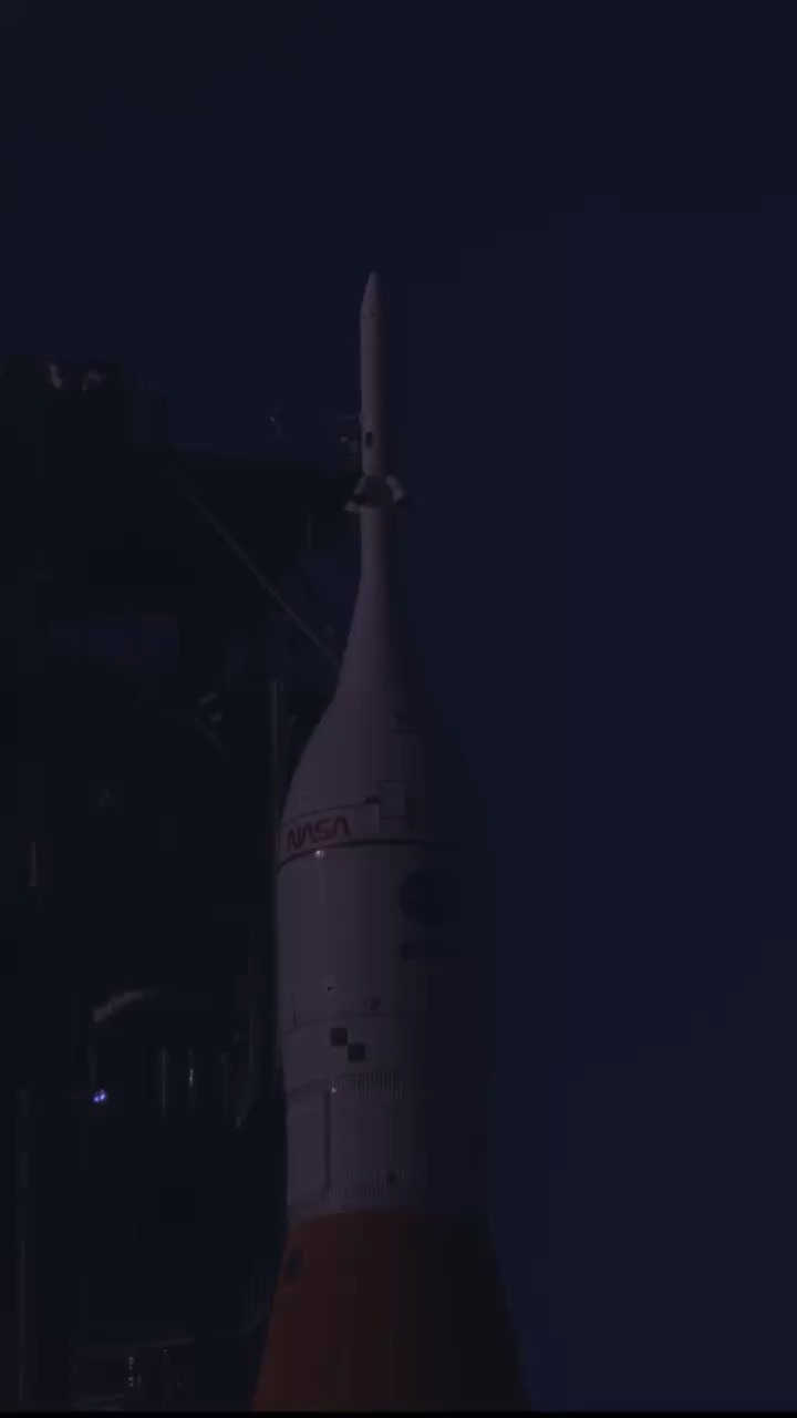 In low blue light, the upper part of a rocket stands beside a dark tower structure: a white conical crew section carrying the red NASA wordmark, topped by a slender launch abort tower, with an orange stage beginning at the bottom of the frame.