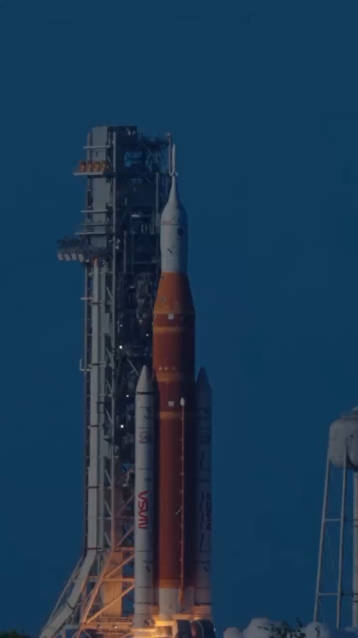 An orange core stage flanked by two white solid rocket boosters stands beside a grey launch tower against a deep blue twilight sky, with a white crew capsule and escape tower at the top. A red NASA worm logo is painted on the near booster.