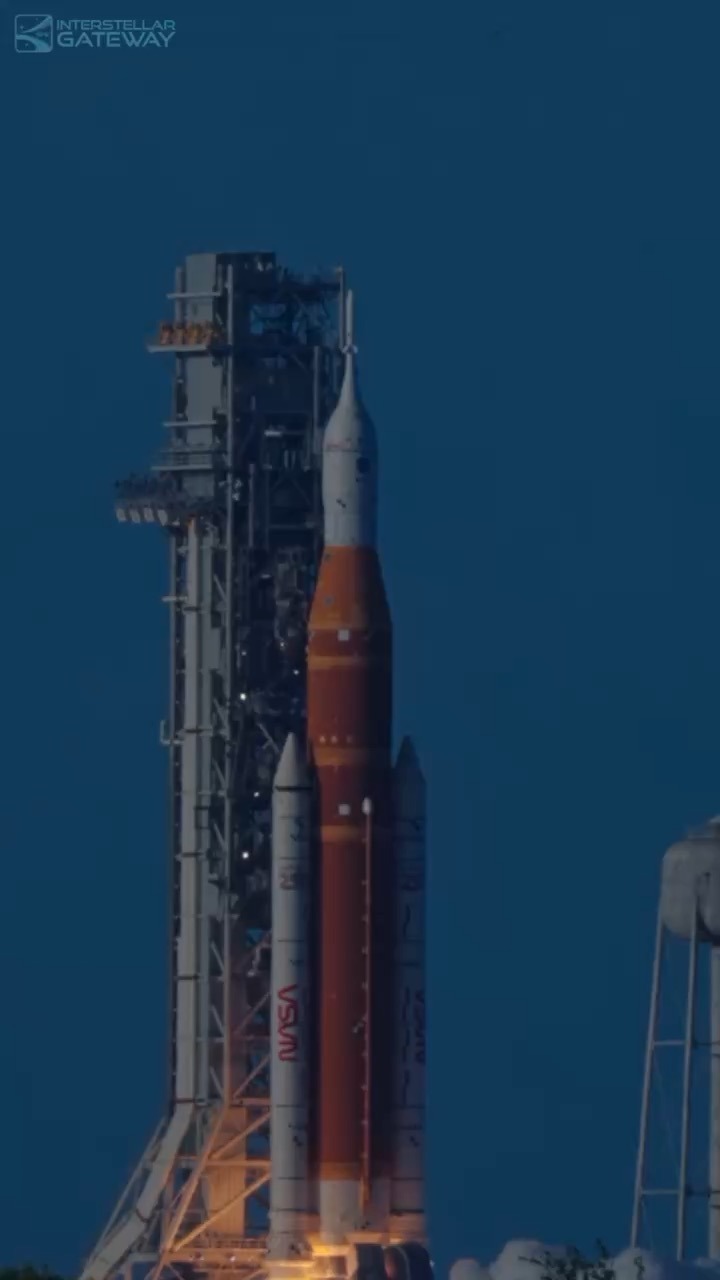 An orange core stage with two white solid rocket boosters stands next to a tall grey launch tower in deep blue twilight, a white crew capsule and escape tower on top and a red NASA worm logo on the near booster.