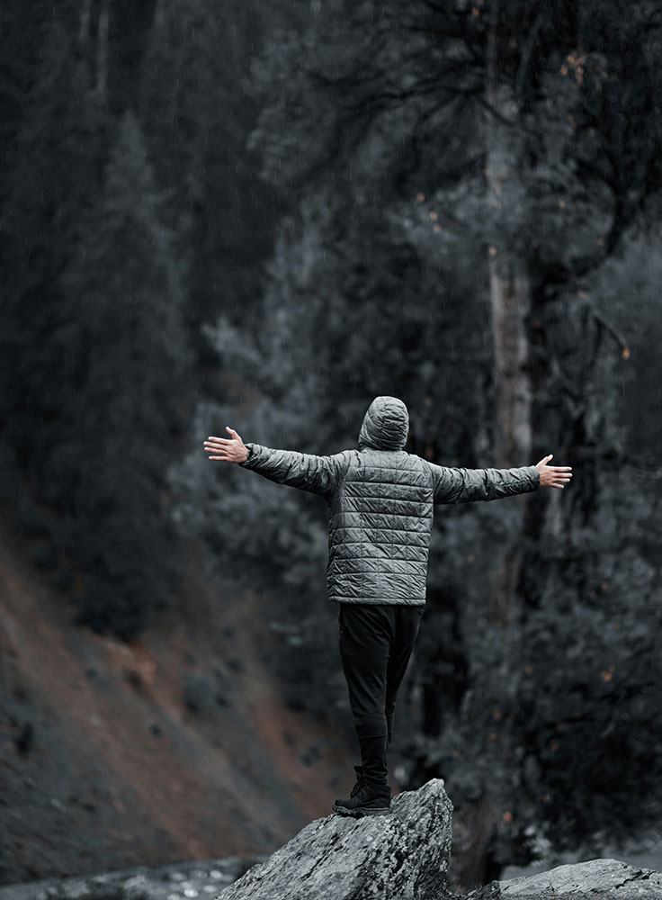 Person mit Kapuzenjacke steht mit ausgebreiteten Armen auf einem Felsen, umgeben von Natur – Symbol für Durchhaltevermögen.