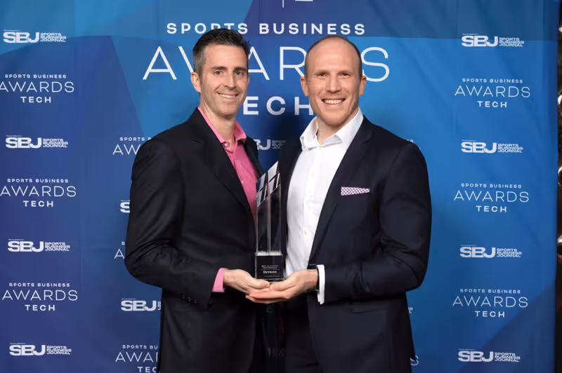 Two men in suits smiling and holding a tall rectangular award trophy in front of a blue backdrop with 'Sports Business Awards Tech' and 'SBJ Sports Business Journal' logos.