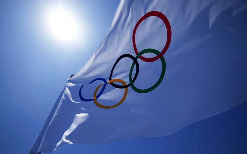 White Olympic flag waving against a clear blue sky with the sun shining.