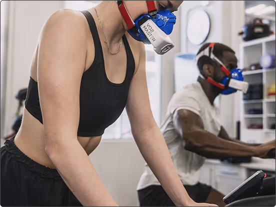 Two people exercising indoors wearing VO2 Master respiratory masks on stationary bikes.