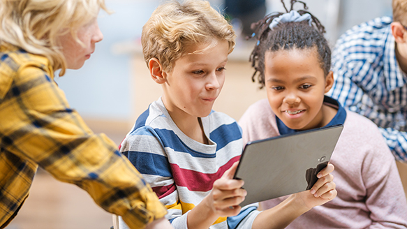 A photo of a diverse group of three children excitedly looking at an ipad in a classroom setting.