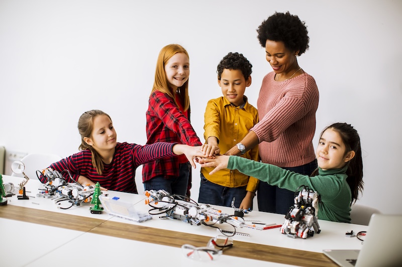A teacher and a group of four diverse students, standing over a robotics STEM project and placing their hands on each other symbolizing team working.