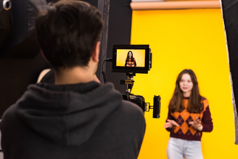 A photo of a male holding a professional video camers, in the screen you see a young women in front of a yellow background