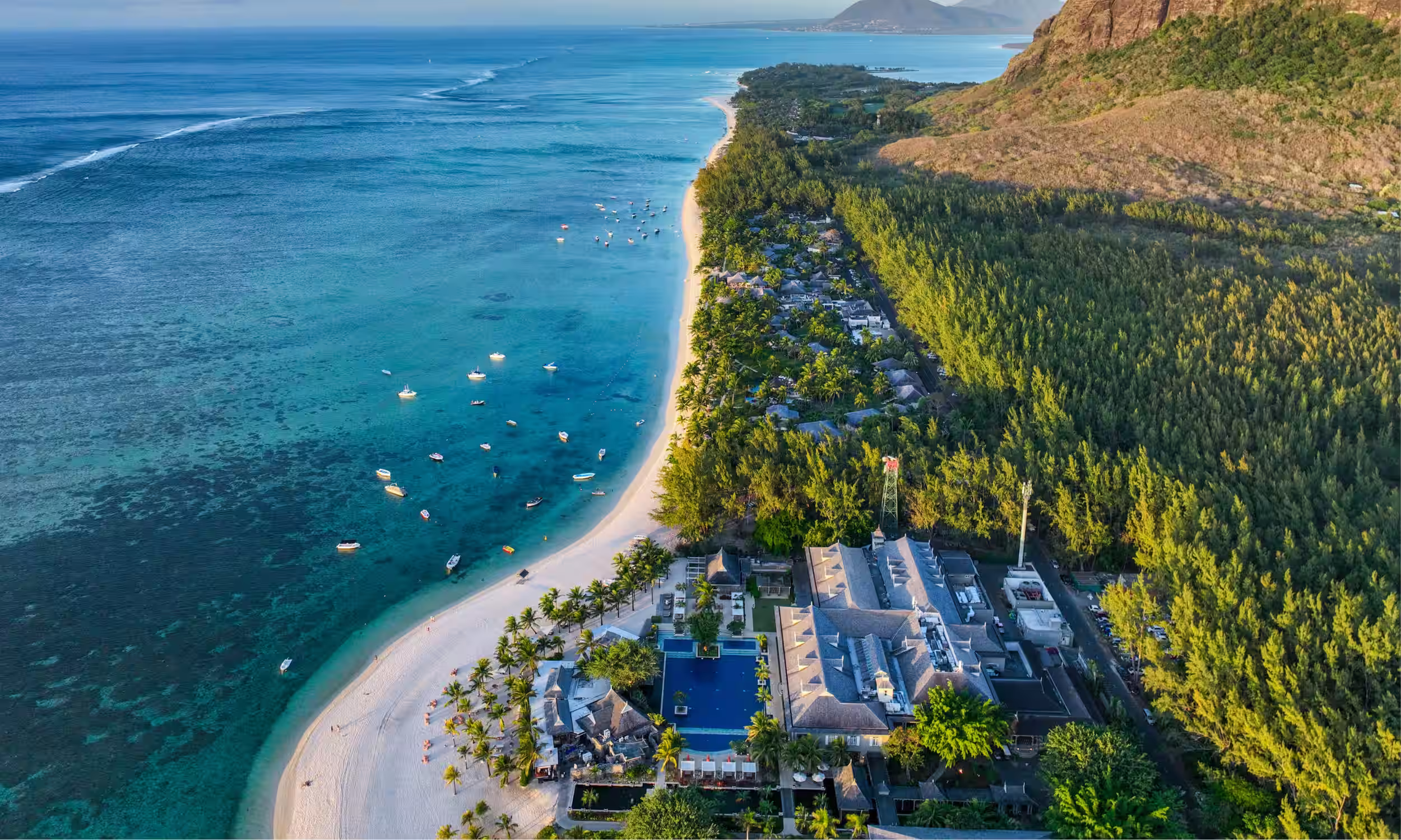 Aerial view of Le Morne Peninsula in Mauritius, with turquoise lagoon waters, white sandy beach, boats anchored offshore, lush forest, and the iconic Le Morne mountain rising in the background.