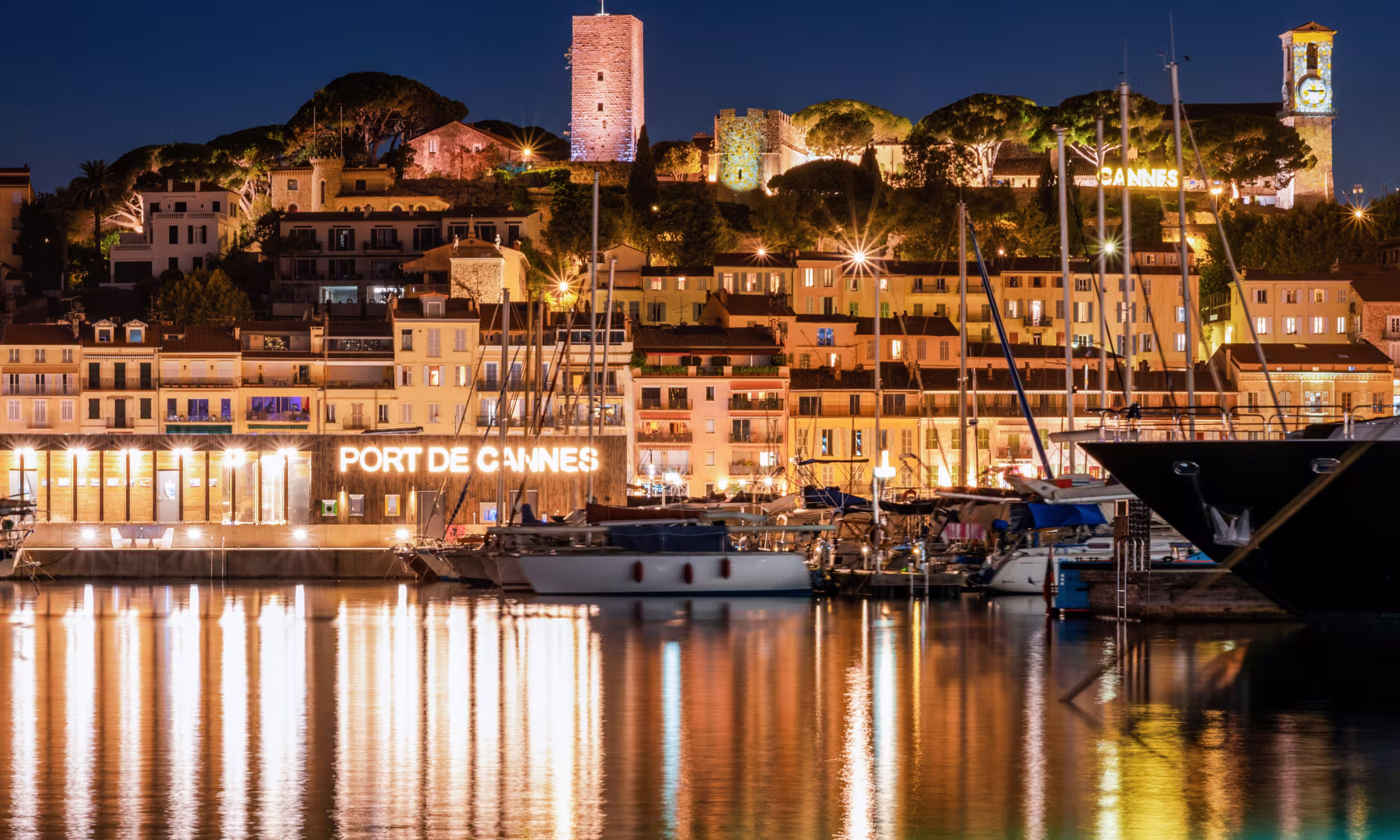 The Cannes harbor at night, with yachts and buildings reflected on the calm sea. 