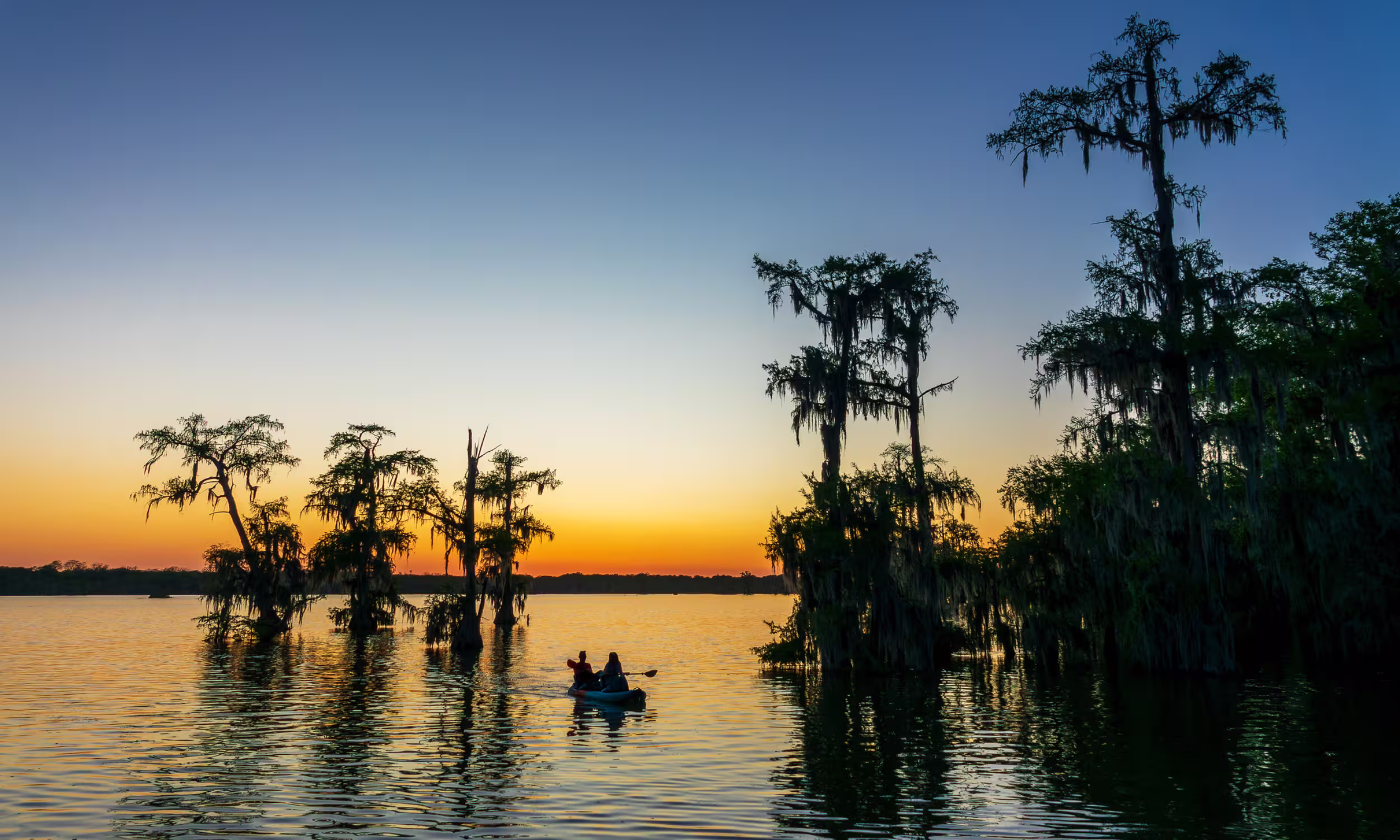 Couple in a canoe at Lake Martin at sunset surrounded by cypress trees and reflections.