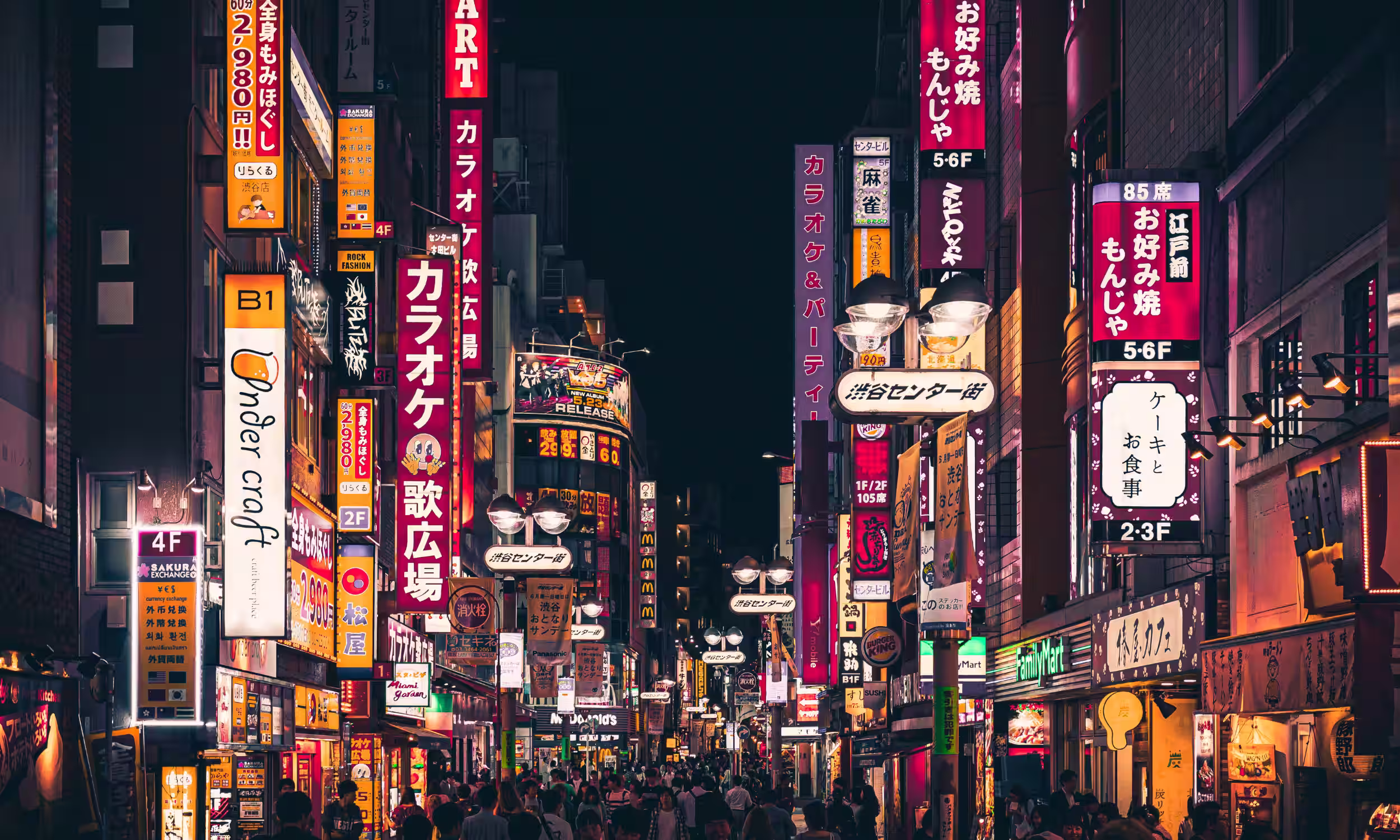 People walking along a brightly lit Tokyo street at night surrounded by neon signs and city buildings.