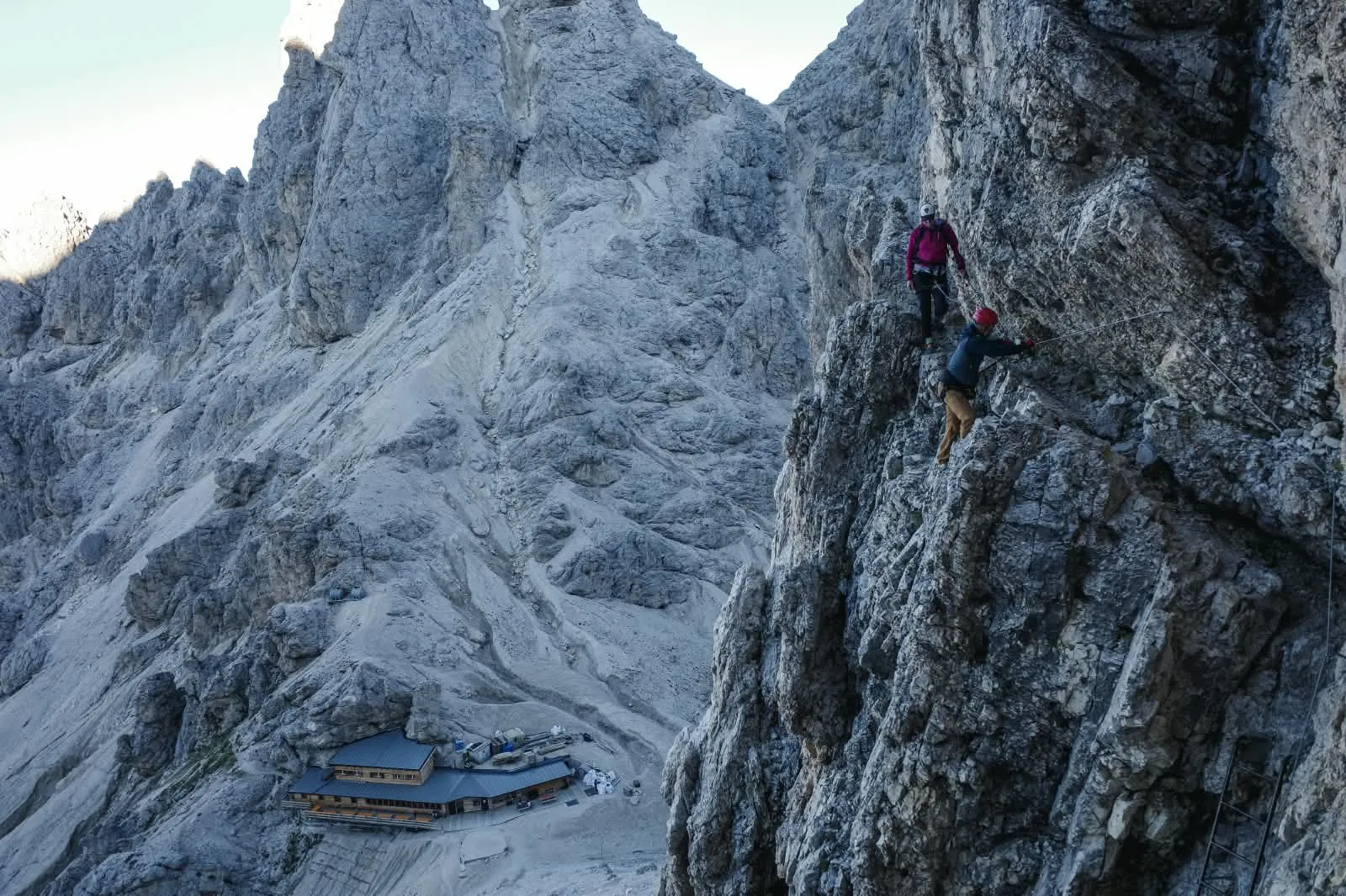 Two climbers on a via ferrata on a steep rock face above a modern mountain hut