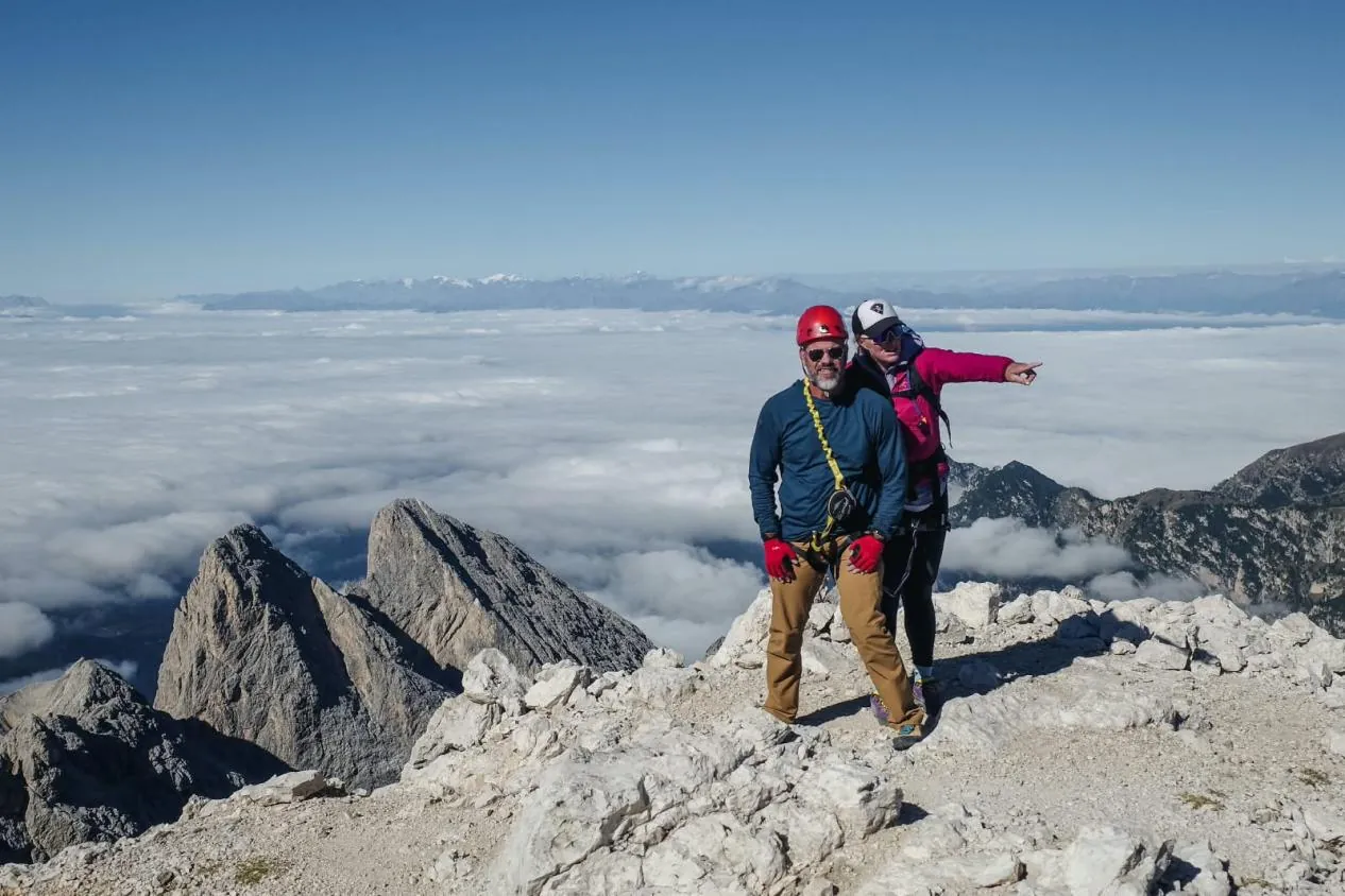 Two climbers in helmets on a summit above a sea of clouds, one pointing toward distant peaks