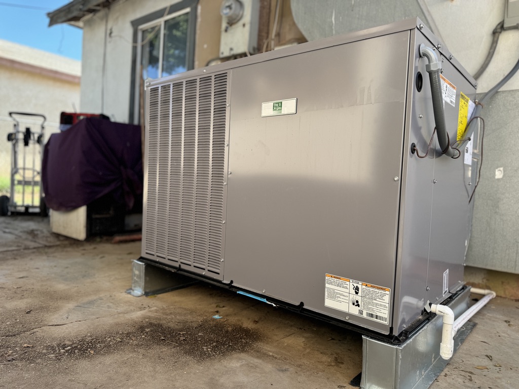 Large gray HVAC unit outside a house on a concrete pad.  Nearby, a dolly and covered equipment. 