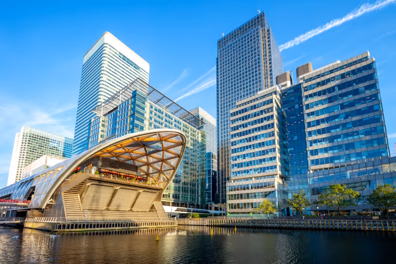 Modern glass office buildings in Canary Wharf reflected in calm water under a blue sky.