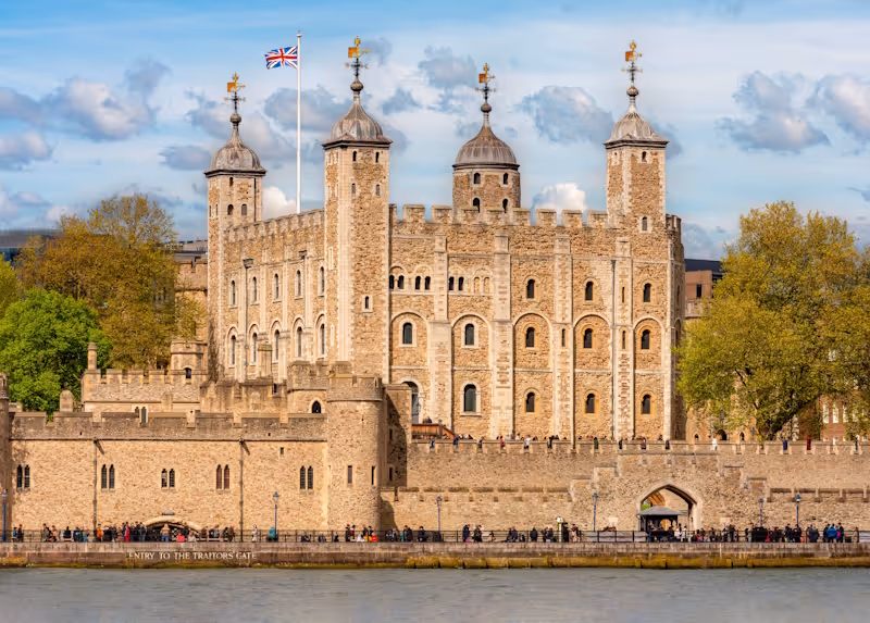 The Tower of London, a historic stone fortress with four tall turrets and a British flag flying atop, beside the River Thames on a partly cloudy day.