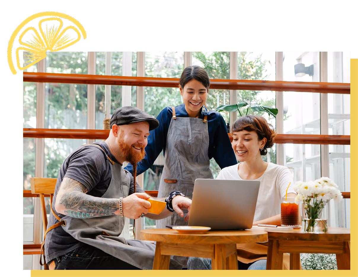Three smiling people in aprons gathered around a laptop in a bright cafe, with a cup of coffee and flowers on the table.