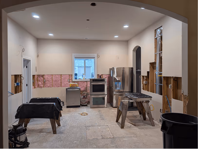 Kitchen stripped to bare walls and subfloor with new recessed lighting rough-ins visible in the ceiling