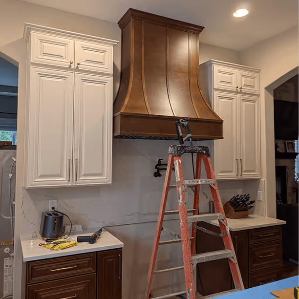 Copper bell-shaped range hood being installed above the range between white upper cabinets
