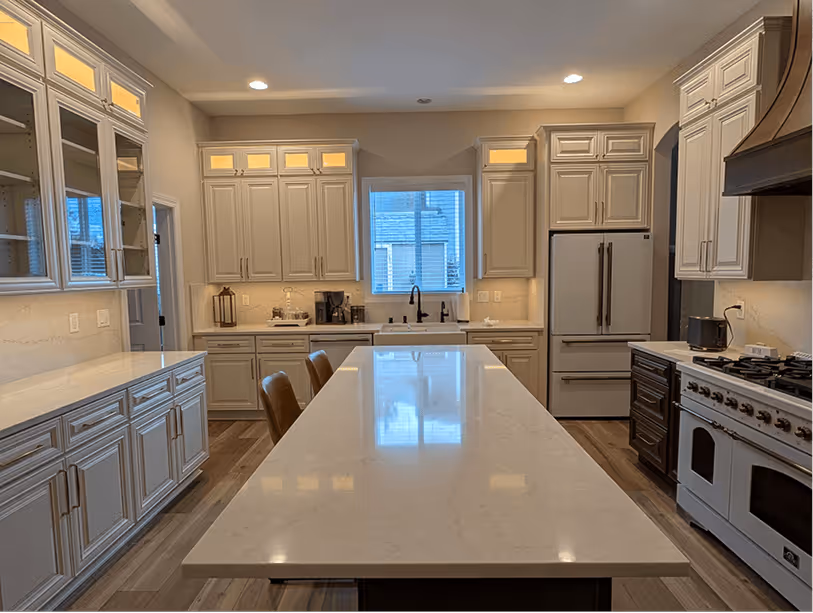 Finished kitchen looking toward t ahe sink wall, white quartz island with bar stools in foreground, lit glass-door cabinets behind