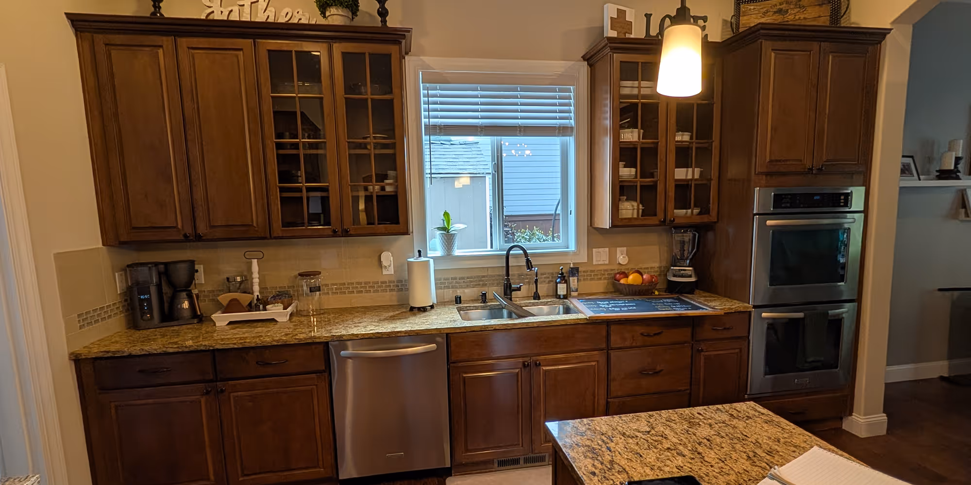 An outdated kitchen with dark brown wooden cabinets, speckled granite countertops, a stainless steel dishwasher, and double wall ovens.