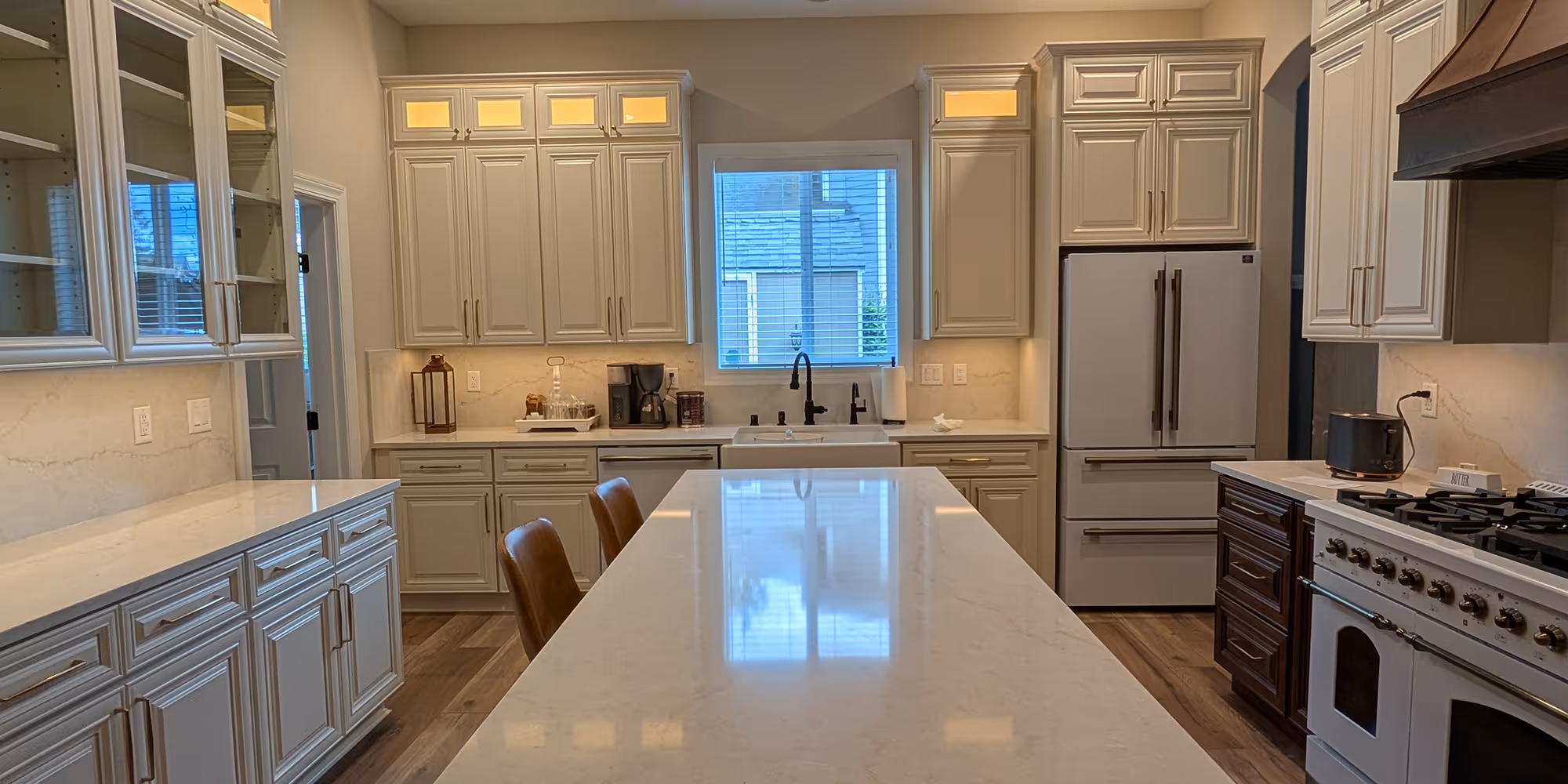 A sleek kitchen with bright white quartz countertops on a central island, off-white cabinetry, and a window above a farmhouse sink.