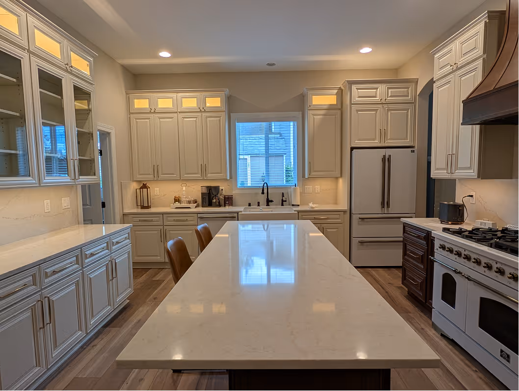 Finished kitchen looking toward t ahe sink wall, white quartz island with bar stools in foreground, lit glass-door cabinets behind