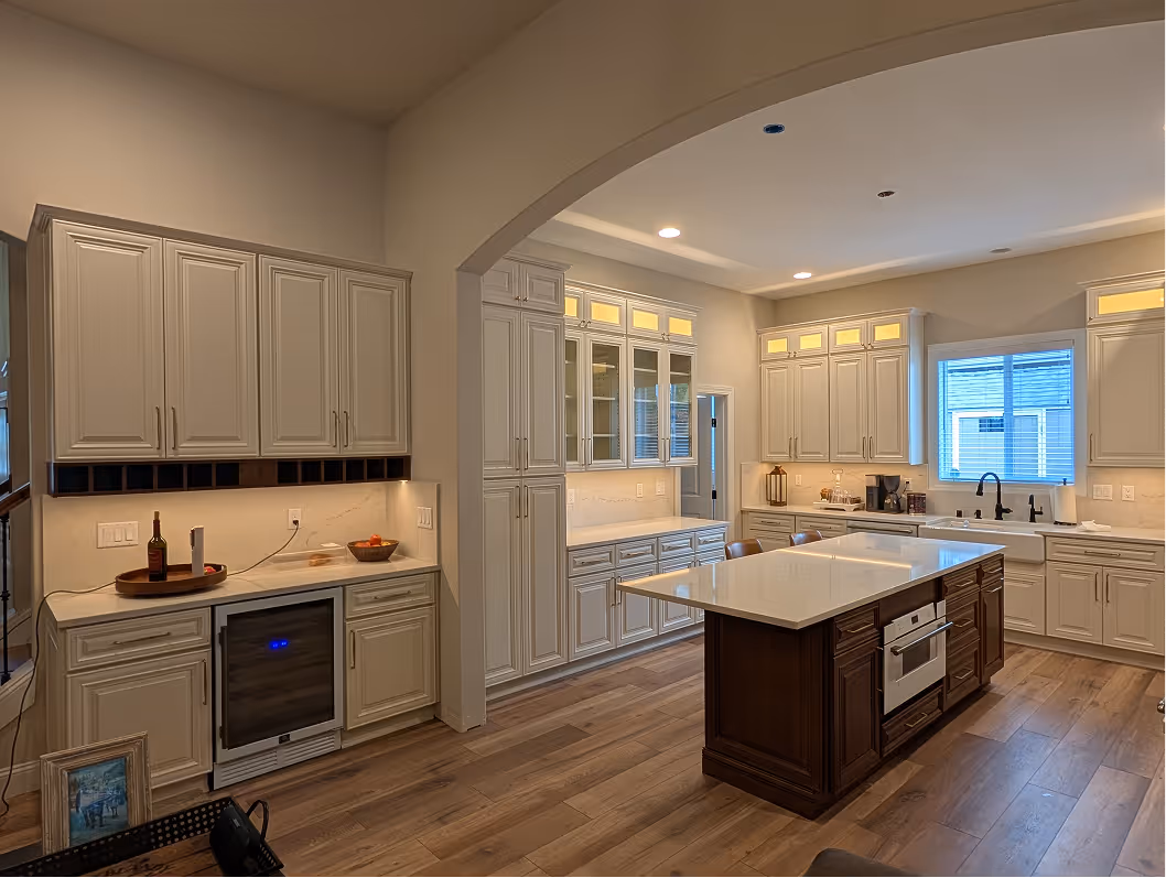 View through an archway of a remodeled kitchen with off-white cabinets, a beverage station with a wine fridge, and a dark wood island.