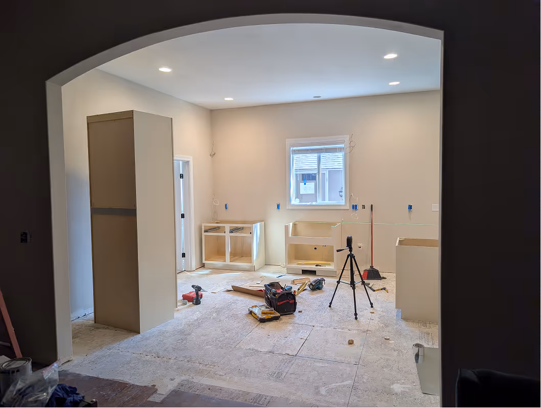 Construction shot of a kitchen remodel with unfinished floors, tools, a laser level on a tripod, and partially installed white cabinet bases.