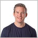 Smiling man with short blond hair wearing a dark blue shirt against a plain background.
