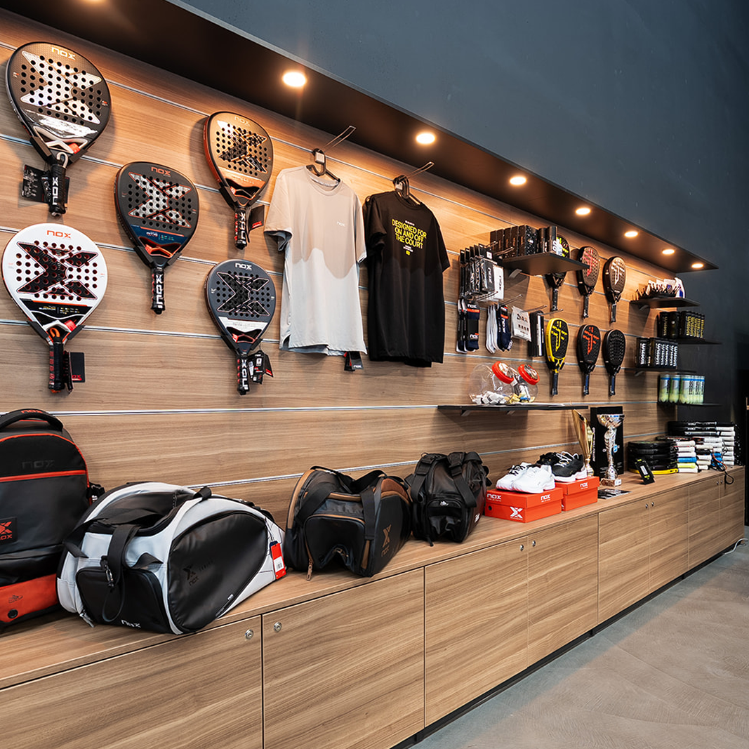 Prodavaonica sportske opreme s padel reketima, torbama, tenisicama i majicama na drvenim policama s osvjetljenjem.