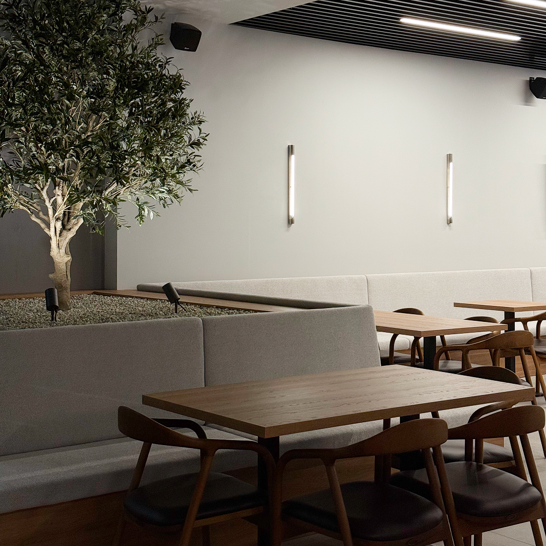 Moderan kafić s drvenim stolovima, sivim klupama i ukrasnim stablom u unutarnjem prostoru s laganim zidnim svjetlima.