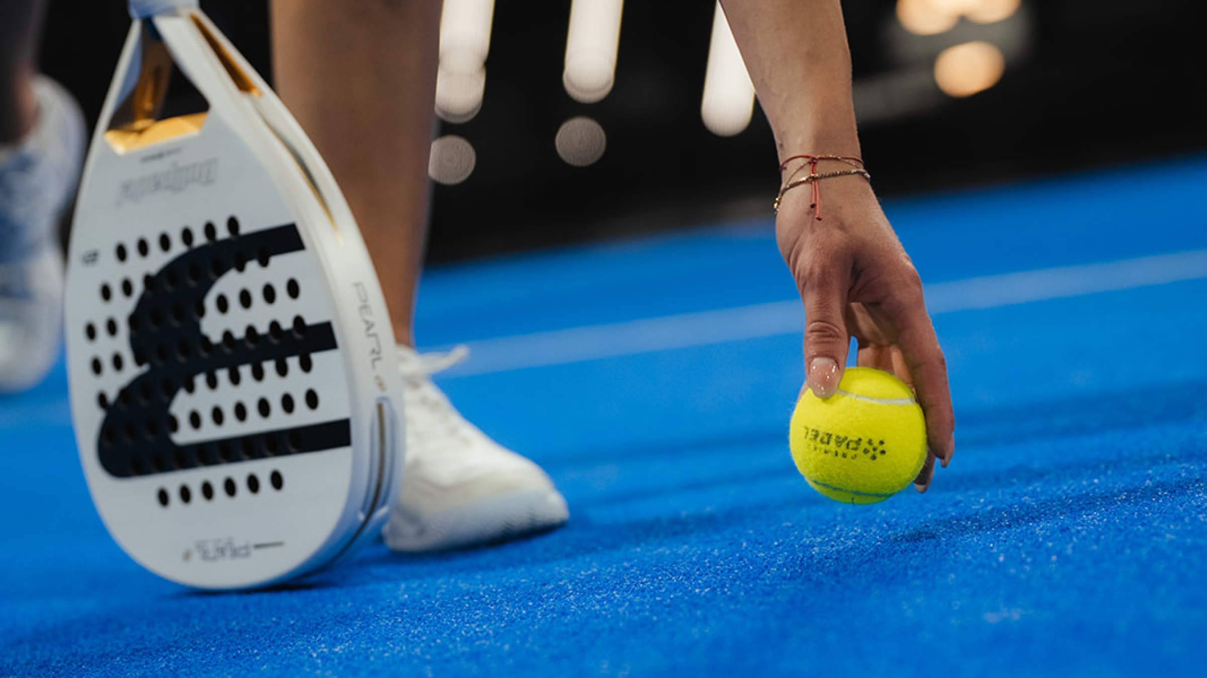 Ruka osobe koja se pruža da podigne žuti padel lopticu s plavog igrališta uz bijelu padel palicu i bijelu tenisicu u pozadini.