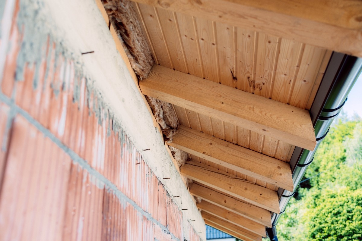 Close-up of roof eaves showing exposed wood rot and dry rot damage along the fascia area above a brick wall, near a gutter system