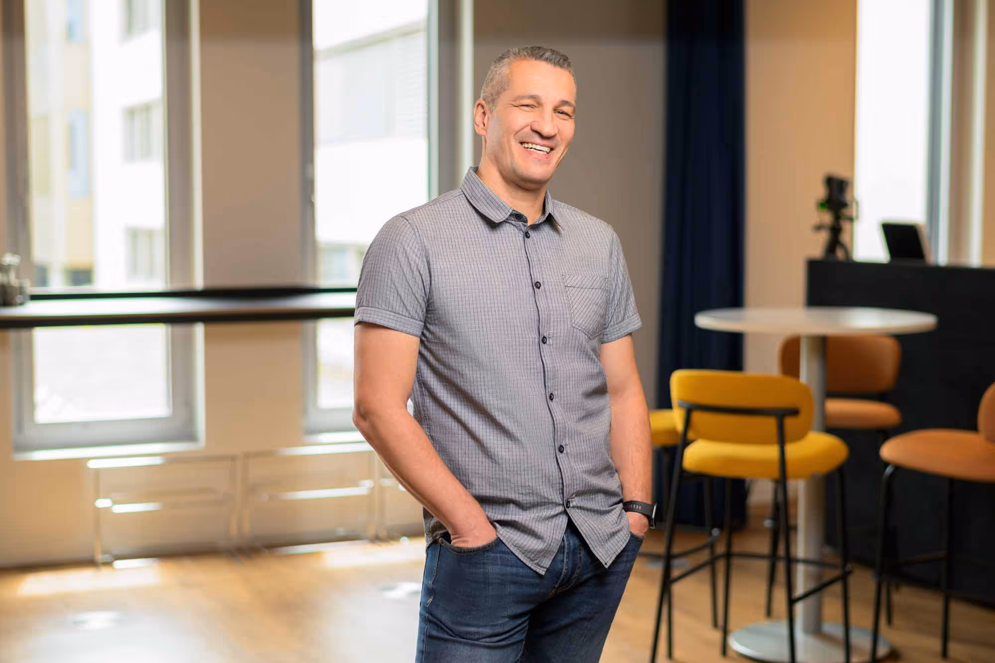 A man in a gray shirt and blue jeans stands in front of a window.