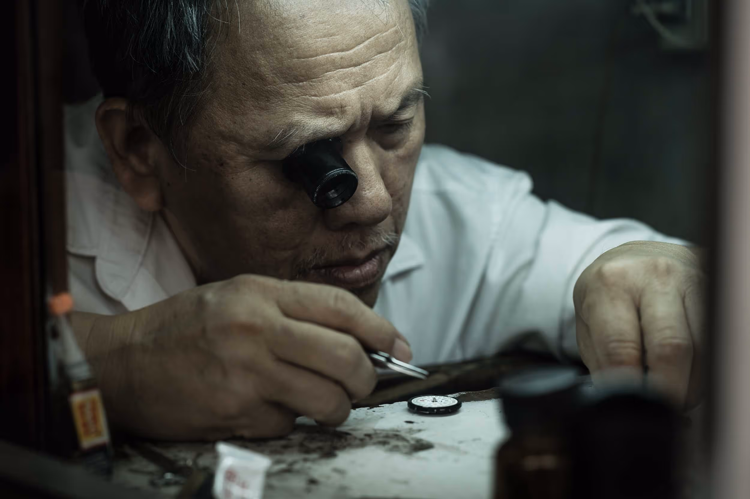 A Thai watchmaker wearing glasses and a white shirt works on a watch.