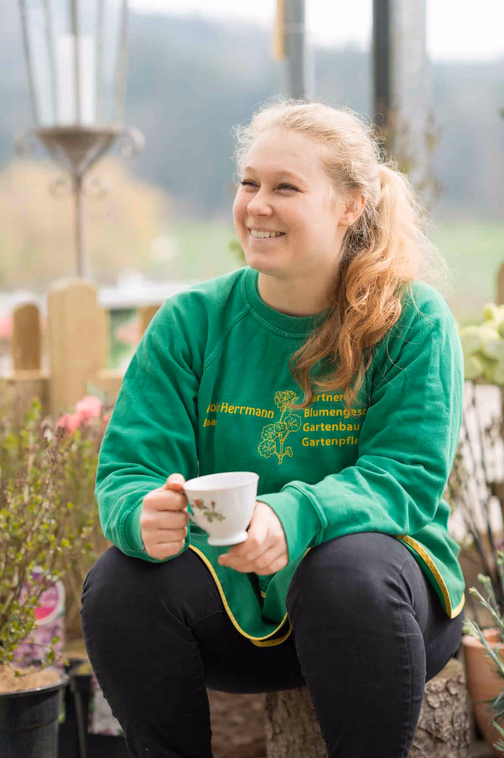 Eine Frau sitzt mit einem grünen Pullover und einer Tasse Kaffee auf einem Baumstumpf.