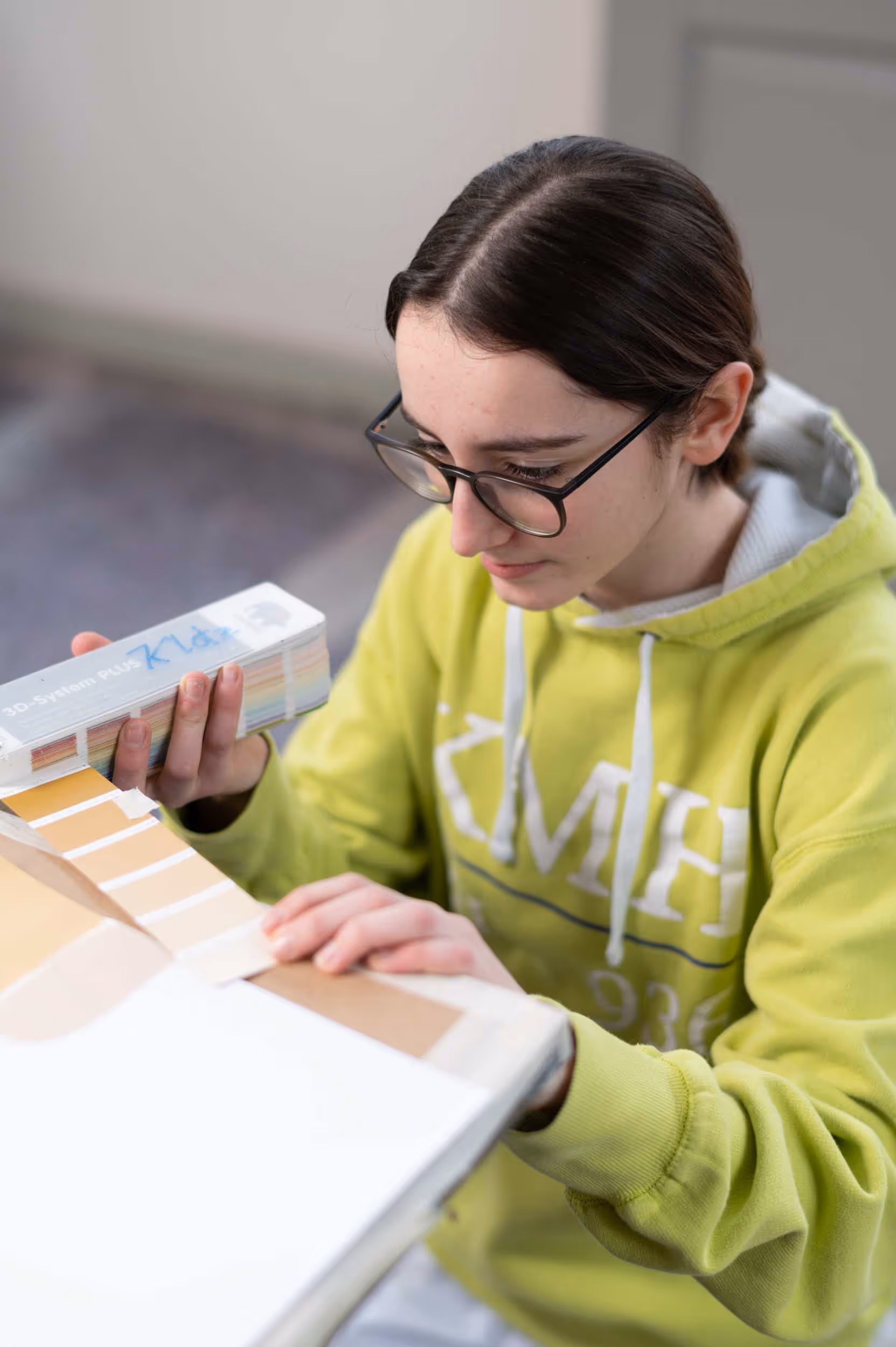A woman with glasses and a green hoodie holds a color chart in her hand.